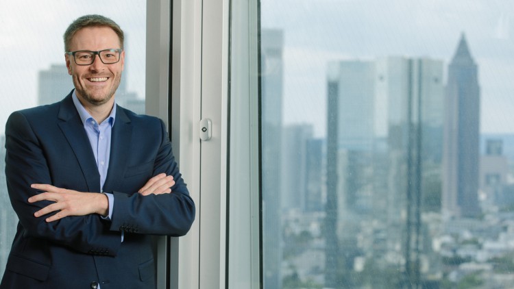 Alexander Schuh steht mit verschränkten Armen vor einem Fenster mit der Frankfurter Skyline (Messeturm) im Hintergrund.