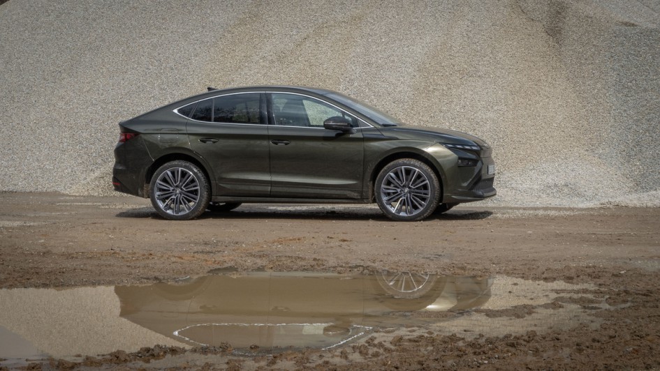 Dunkelgrün lackierter Skoda Enyaq Facelift in der Sonne vor einem Haufen Kieselsteine stehend mit Spiegelung in der davor befindlichen Pfütze