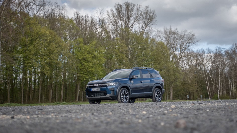 Dacia Bigster Hybrid 155 schräg von vorn auf einem Parkplatz fotografiert. Im Hintergrund sind grün blühende Bäume zu sehen