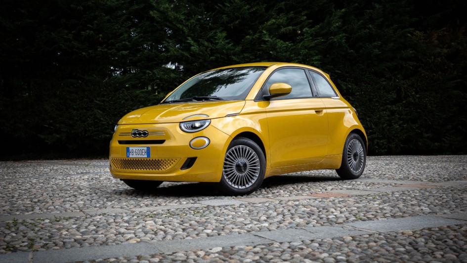 Aufnahme des kompletten Fiat 500 Hybrid in goldgelb schräg von vorn vor Bäumen stehend
